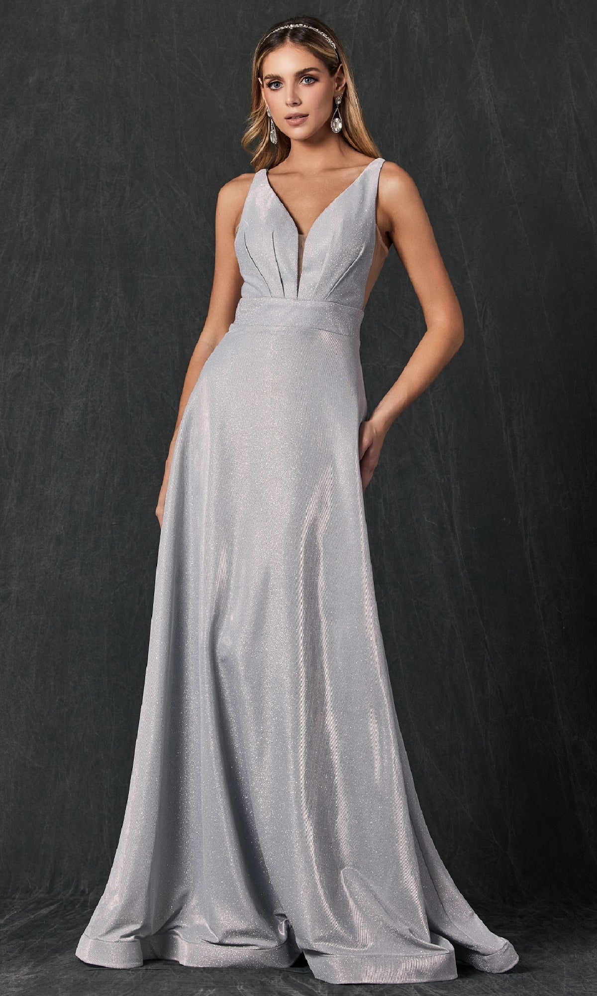 A woman wearing the Juliet 208 Long Prom Dress—a silver, floor-length gown with a sleeveless design and deep V-neckline—stands against a dark textured background.